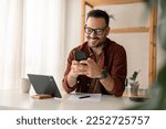 Happy smiling businessman wearing casual clothes and using modern smartphone in his home office, successful employer sitting at desk typing on mobile phone, also using digital tablet, pen and paper.