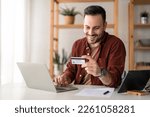 Small photo of Young businessman millennial man consumer working on laptop holding credit card sitting at desk at home doing online banking transaction. E-commerce virtual shopping, secure mobile banking concept.