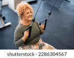 Young woman gym client with curly hair wearing sportswear motivated to exercise focused on her goal, doing cable pulldowns using cable pulley machine to workout her back and arms in gym fitness center