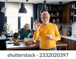 Small photo of Happy confident senior man looking at camera and smiling showing with hand ok sign satisfied with breakfast porridge, standing in front of family members, senior woman and two kids in the kitchen.