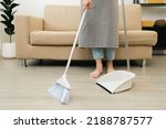 Small photo of Close up housewife using broom and dustpan wearing an apron to clean the living room at apartment. Young woman is happy to clean home. Maid cleaning service.