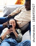 Small photo of Overhead shot of young mixed ethnicity couple relaxing on sofa at home with man watching tv whilst woman lies across lap and checks social media on mobile phone