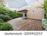 Terrace of a ground floor house with hedges and ornamental plants, slatted acacia hardwood floors twin wooden sunbeds and white awnings
