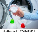 Washing powder detergent, in the background an open washing machine with white laundry. Hand holds a measuring cup of washing powder.
