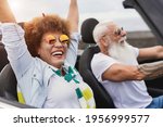 Small photo of Trendy senior couple having fun inside convertible car - Multiracial mature people on a road trip with cabriolet car