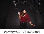 Small photo of Female magician makes with soap bubbles show, an illusionist in theatrical clothes, on black background. Woman actress in stage costume. Concept of theatrical performance and fun show. Copy space