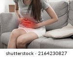 Small photo of asian young woman suffering stomach ache sitting on couch in living room at home, people painful stomachache, gynecology, menstrual pain , medical and health care concept