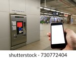 Small photo of hand holding white blank screen mobile phone with self service ticket or ticket vending machine at train station, transportation, technology, internet, network connection and social media concept