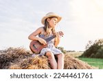 Small photo of Pretty little girl in panama hat sit at top of haystack looking away and play ukulele. Carefree child resting on hayrick. Outdoor walking. Beautiful sunny sky. Summer vacation, countryside lifestyle.