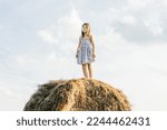 Small photo of Pretty little girl stand at top of haystack looking away. Full length photo, from below view. Resting on hayrick. Outdoor walking. Beautiful blue sky. Freedom. Summer vacation, countryside lifestyle.