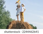 Small photo of Little girl and boy together launch yellow toy airplane standing at top of haystack. Children on hayrick from below view. Freedom. Beautiful blue sky. Summer vacation, countryside lifestyle.