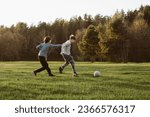 Small photo of Foxy teenager in gray grabbing friend on clothes attempting to overtake boy throughout running towards white ball. Competitive teens playing football on green field near forest and enjoying game.