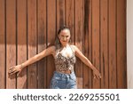 Small photo of Young and beautiful woman, with ponytail, wearing a floral top and jeans, serious and angry with open arms leaning on a wooden door. Concept beauty, seriousness, anger, reproach.