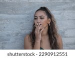 Small photo of Latina and Hispanic girl, young and nonconformist, rebellious, smoking a cigarette rolled by herself, on a gray background. Concept tobacco, smoke, fire, addiction, gangs.