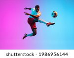 Jumping, flying. One young African man, professional soccer football player training isolated on gradient blue pink background in neon light. Concept of action, energy, sport. Copy space for ad