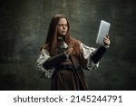 Small photo of Shock. Vintage portrait of young beautiful girl in image of medieval warlike woman using modern gadget isolated over dark background. Comparison of eras, history, renaissance style. Fashion, style