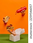 Small photo of Food pop art photography. Female hand sticking out orange paper with ketchup over burger on hand sticking out food box. Concept of taste, creativity, art. Complementary colors. Copy space for ad, text