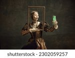 Small photo of Portrait of pretty young girl, royal person in vintage dress holding picture frame and taking selfie with phone against dark green background. Concept of history, renaissance art, comparison of eras