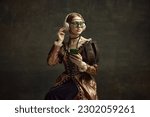 Portrait of young beautiful girl in vintage dress, trendy sunglasses, headphones and mobile phone against dark green background. Concept of history, renaissance art, comparison of eras, social media