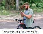 Happy bearded man in jeans clothes stand near bicycle bike on sidewalk in green park outdoors use mobile cell phone chat online. People active urban healthy lifestyle cycling concept