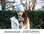 pretty young woman with curly brown hair wears a woollen hat for the cold and covers her face with a dry leaf from a tree. Autumn and cold concept.