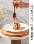 Small photo of Woman pouring caramel syrup onto piece of layered honey cake on white plate. Drip of caramel in a spoon. Holiday cake. Celebrations food concept. Slice of birthday honey cake decorated with caramel