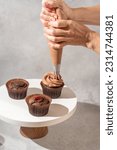 Small photo of Tasty cupcakes with sweet whipped chocolate frosting on light background. The confectioner’s hands decorating cupcakes with chocolate cream by pastry bag. The process of decorating cupcakes