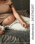 Small photo of Close-up of young woman with pregnant belly. Attractive pregnant woman with large tummy sitting on the bed and waiting for a newborn baby. Concept of pregnancy, motherhood, procreation, expectation