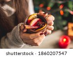 Woman in a warm sweater is holding in her hands a cup of aromatic hot mulled wine on the background of a Christmas tree with lights. Concept of a festive atmosphere and cozy winter mood. Closeup