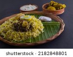 Small photo of Kolkata style mutton biryani with potato and egg served on clay plate and banana leaf with mutton curry and curd raita. shot against black background.