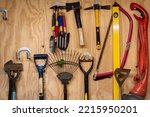 Small photo of Assortment of DIY gardening tools and equipment hanging organised on wooden wall inside garden shed. Tools include rake, shovel, hammer, fork, trowel, spirit level measure, saw, axe, hatchet etc.