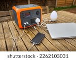 Small photo of Portable power station solar electricity generator outdoors on wooden table with laptop, mobile phone and lamp electronic devices charging. Wireless charging lithium battery backup for use off grid.