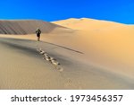 Man wearing straw hat, blue long sleeve shirt, shorts, and daypack walks up pristine sand mountain leaving footprints at Kelso Dunes California, sunny blue sky day