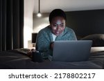 Smiling black woman, holding cup of coffee, lying on bed, looking at laptop screen