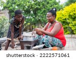 Small photo of image of african mother and her daughter in local kitchen- black woman stirring a sauce pan and a girl looking into the sauce pan- food concept