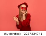 Excited stylish happy laughing girl holding hand on face or keeping hand on cheek, wearing beret headwear, red dress with heart glasses and looking up isolated on colored pink background.