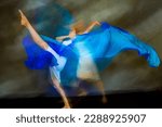 Small photo of Mystical, blurred image of a female professional dancer in a white dress, dancing with a floating, blue, gauzy fabric in a studio shot, against a gray background.