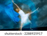 Small photo of Mystical, blurred image of a female professional dancer in a white dress, dancing with a floating, blue, gauzy fabric in a studio shot, against a gray background.