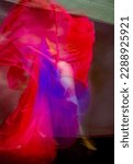 Small photo of Mystical, blurred image of a female professional dancer in a white dress, dancing with floating, red and blue, gauzy fabrics in a studio, shot against a gray background.