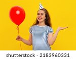 Pretty smiling young woman in a birthday cap gestures with hand, holds red inflated helium balloon, celebrates birthday or special occasion. Holiday and party concept