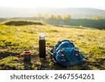 In the peaceful landscape, a backpack, thermos, and mug are placed on a grassy hill surrounded by lush vegetation, vast sky, and majestic mountains in a beautiful highland scenic view