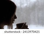 Long hair woman standing among the window and drinking hot tea from mug. Winter landscape. Lady drinks coffee looking at snowfall. Depression seasonal affective disorder female sad alone. Flu season