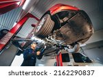 Small photo of Young car mechanic at repair service station inspecting car wheel and suspension detail of lifted automobile. Bottom view.