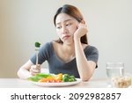 Small photo of Diet in bored, unhappy beautiful asian young woman, girl on dieting, holding fork at broccoli in salad plate, dislike or tired with eat fresh vegetables. Nutrition of clean, healthy food good taste.