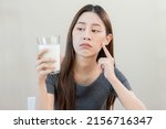 Small photo of Allergy asian young woman, girl looking, holding glass of milk, face in thinking before drink milk as it may upset her stomach ache, pain. Lactose intolerance and dairy food , health problem concept.