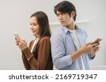 Small photo of Infidelity, suspicion asian young couple love standing back to back, husband watching his wife using mobile phone, spying his girlfriend while woman typing a message, man distrust and jealousy at home