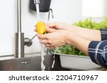 Close up hand of asian young housekeeper woman, washing sweet pepper, yellow paprika, vegetables with splash water in basin of water on sink in kitchen, preparing fresh salad, cooking meal.Health food