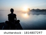 Silhouette of a woman sitting on the pier against the sunset sky with copy space. Loneliness concept. Rising sun over the lake in the fog. Be on your own.