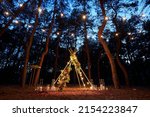 Small photo of Festive string lights illumination on boho tipi arch decor on outdoor wedding ceremony venue in pine forest at night. Vintage string lights bulb garlands shining above chairs at summer rural wedding.