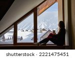 Small photo of Woman being cold sits on windowsill, warming up with tea mug, looking at snow covered mountains. Female drinks coffee in cozy chalet wearing thermal underwear layer. Rising heating bills on winter.